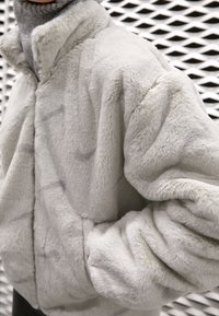 Světle šedá umělá kožešinová bunda s vysokým límcem, dlouhými rukávy a bočními kapsami, která se vyznačuje měkkým, texturovaným povrchem a volným střihem.