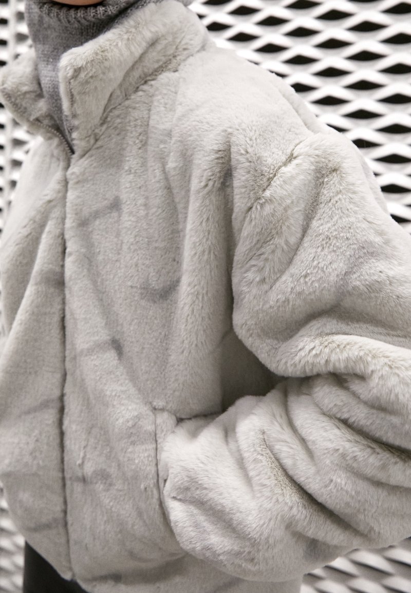 Světle šedá umělá kožešinová bunda s vysokým límcem, dlouhými rukávy a bočními kapsami, která se vyznačuje měkkým, texturovaným povrchem a volným střihem.