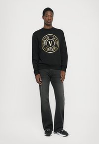 Črn pulover s krožnim zlatim logotipom, ki vsebuje "VERSACE JEANS COUTURE." Oblečen z temnimi kavbojkami in črnimi športnimi copati. Gladka tekstura, sproščen kroj.