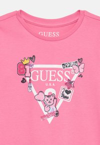 Rózsaszín pamut póló, háromszög alakú grafikával, amelyen a "GUESS" felirat, egy medve illusztráció, szívek és csillogó elemek találhatók.