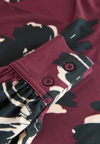 Dlhý rukáv v burgundskej farbe s čiernym a krémovým kvetinovým vzorom. Obsahuje manžetu s skladmi a dva fialové gombíky na zapínanie.