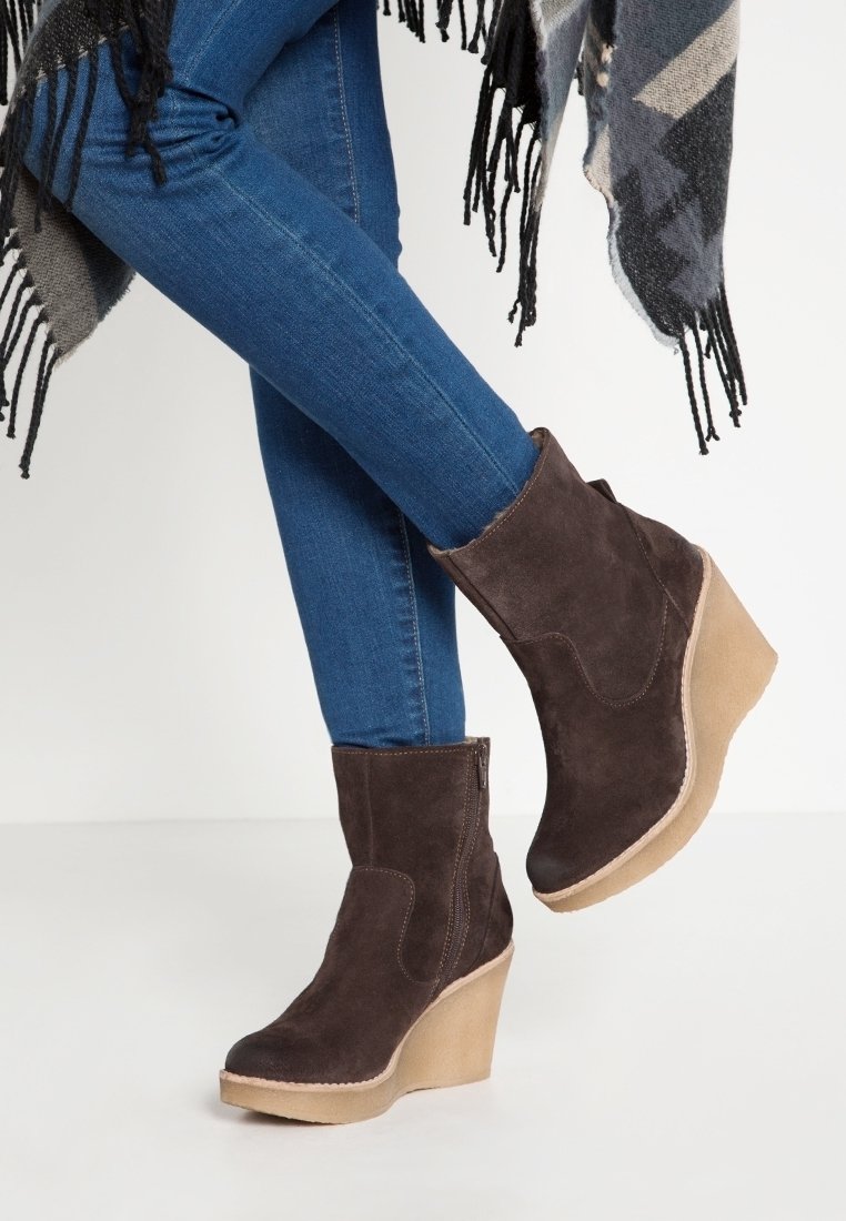 Brown suede wedge ankle boots with a textured finish, featuring a side zipper and a contrasting tan platform sole, worn with blue jeans.