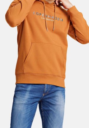 Mand justerer hætten på en brændt orange sweatshirt med teksten "Copenhagen LINDBERGH", kombineret med falmede blå jeans mod en hvid baggrund.
