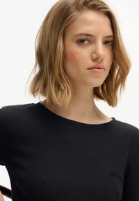 Chemise noire à manches longues côtelée avec un col rond et un logo brodé discret sur la poitrine. Texture lisse, coupe ajustée.