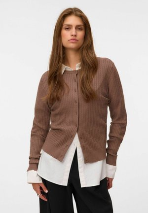 Femme aux longs cheveux bruns porte un cardigan marron boutonné sur une chemise blanche et un pantalon noir sur un fond clair uni.