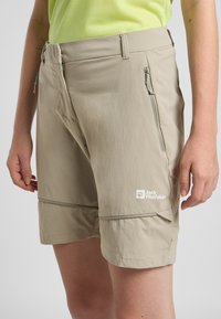 Pantaloni corti beige realizzati in un materiale leggero, con tasche con zip e un logo discreto. Gli accenti includono una cucitura ricamata vicino all'orlo.