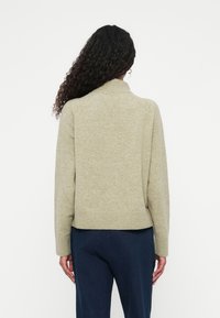 Suéter tricotado verde claro con un cuello alto, puños y dobladillo acanalados, que presenta un corte relajado. Lleva puesto un pantalón azul oscuro.