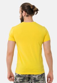 Gelbes T-Shirt mit kurzen Ärmeln aus Baumwolle, mit Rundhalsausschnitt und figurnaher Passform. Kombiniert mit Camouflage-Shorts.