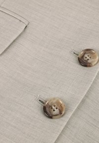 Tejido gris claro con una superficie texturizada que presenta dos botones redondos veteados, cada uno con cuatro agujeros, y un bolsillo lateral.