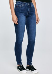 Blugi bleumarin cu croială strâmtă purtați de o persoană în picioare, asortați cu adidași negri cu talpă joasă și un top negru, pe un fundal simplu gri deschis.