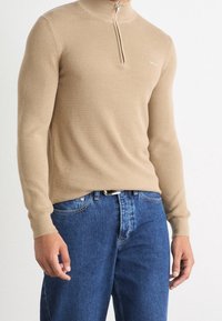 Uomo che indossa un maglione beige a metà zip con texture e jeans di denim blu, in piedi su uno sfondo bianco semplice.