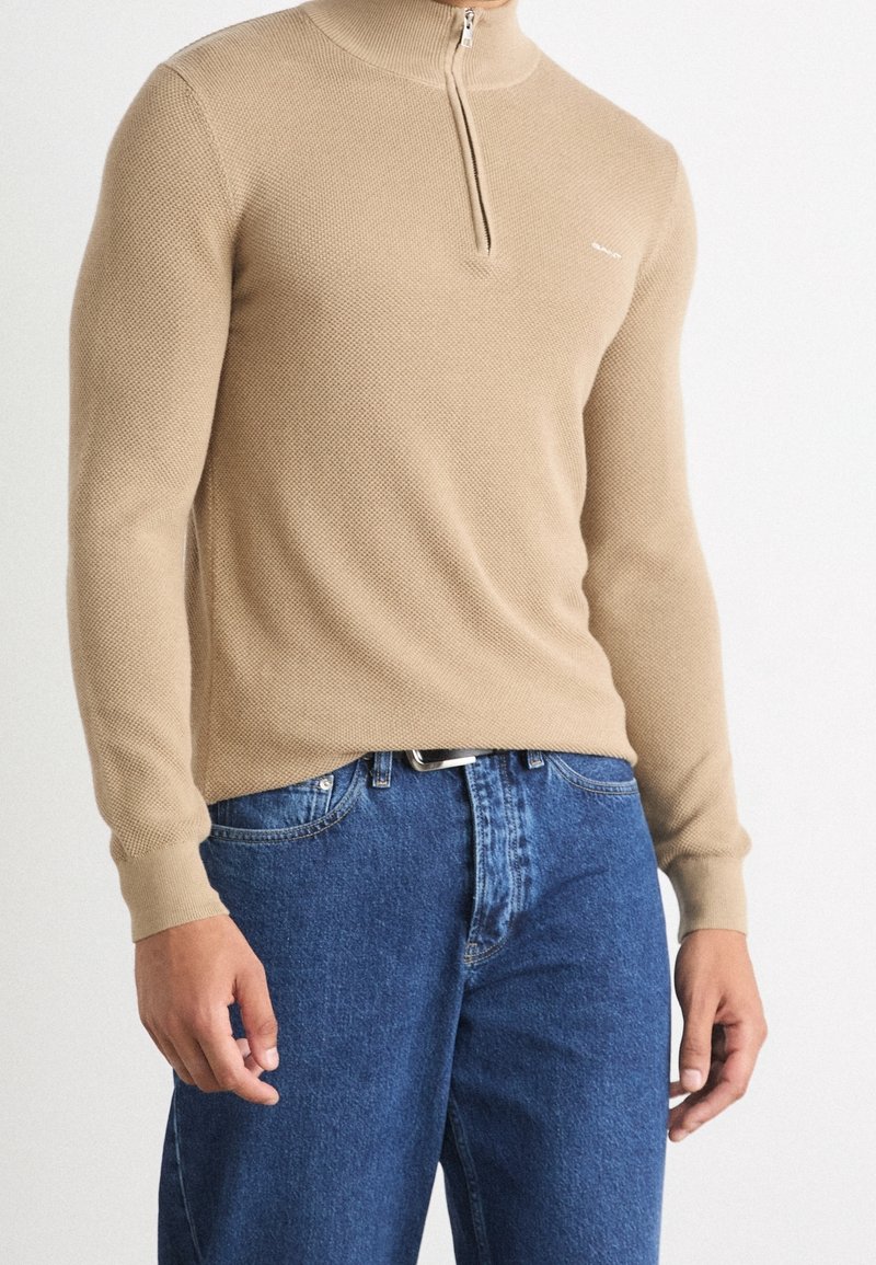 Uomo che indossa un maglione beige a metà zip con texture e jeans di denim blu, in piedi su uno sfondo bianco semplice.