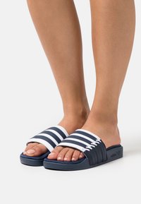 Navy blue slide sandals with white striped straps, a flat design, and textured footbed for grip, featuring subtle branding on the side.