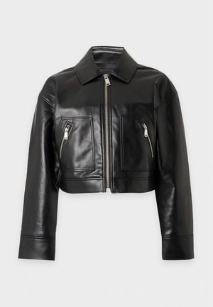 Femme portant une veste en cuir noire, un haut beige, un jean bleu taille haute à jambes larges et des chaussures noires pointues, debout les mains dans les poches.