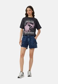 T-shirt graphique noir avec un design rose, associé à un short en denim bleu cargo avec poches latérales et coupe décontractée. Chaussures de sport blanches.