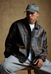 Barna bőrkabát laza fazonban, mellzsebbel és fekete gombokkal, fehér ing és szürke melegítőnadrág felett viselve. Szürke sapka hímezett részlettel.