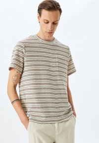 Camiseta ligera a rayas en beige con líneas marrón oscuro, con mangas cortas y un bolsillo en el pecho. Tejido suave y texturizado.