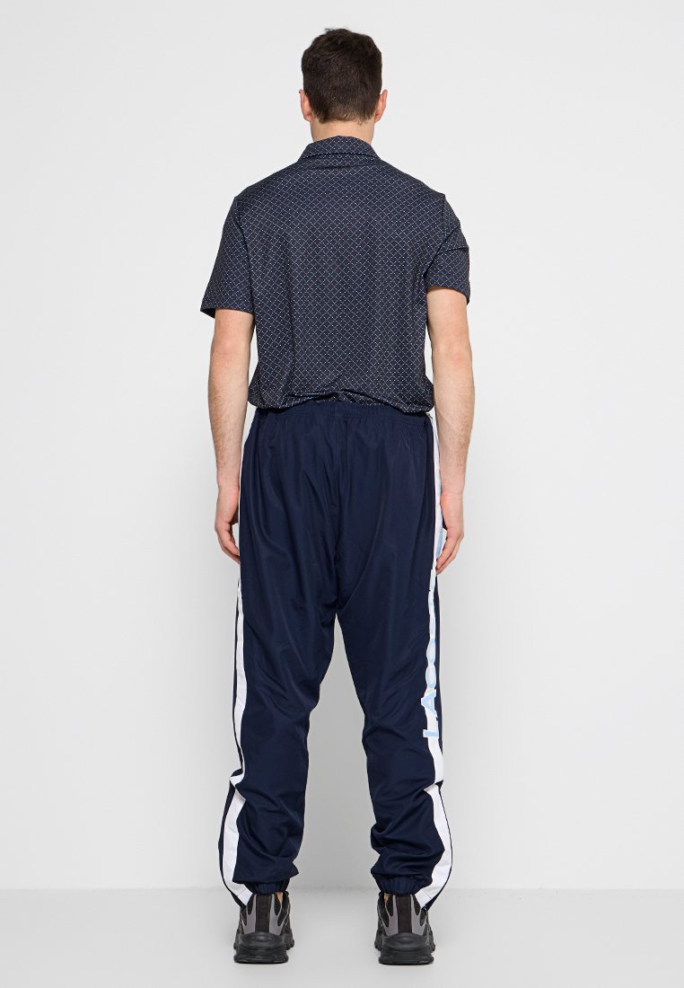 Pantalons de survêtement navy avec des rayures blanches sur les côtés, associés à une chemise à manches courtes à motifs dans des couleurs sombres. Le matériau semble léger et respirant.