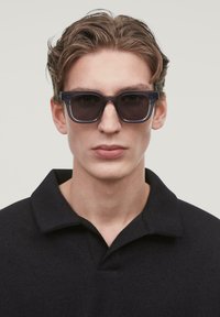 Occhiali da sole con lenti scure e una robusta montatura nera. L'abbigliamento è composto da una maglietta polo nera con colletto classico. Design minimalista.