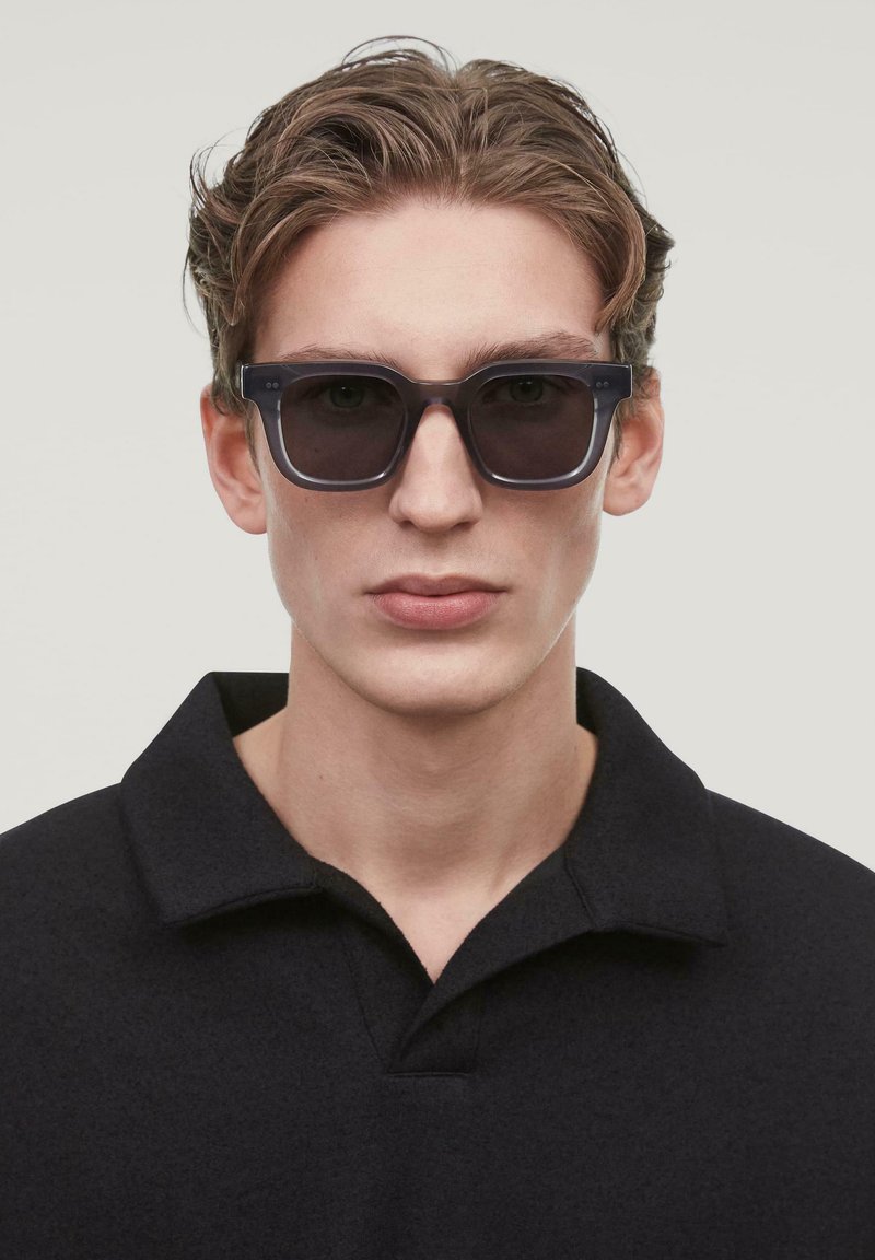 Occhiali da sole con lenti scure e una robusta montatura nera. L'abbigliamento è composto da una maglietta polo nera con colletto classico. Design minimalista.