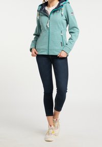 Una chaqueta de forro polar en tono verde azulado con capucha azul marino y bolsillos con cremallera, que cuenta con cordones decorativos y un emblema circular en la manga.