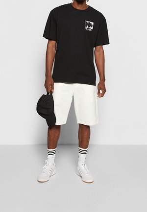 Mężczyzna w czarnej koszulce z nadrukiem, białych spodenkach do kolan, białych trampkach, paskowanych skarpetkach, trzymający czarną czapkę w jednej ręce.