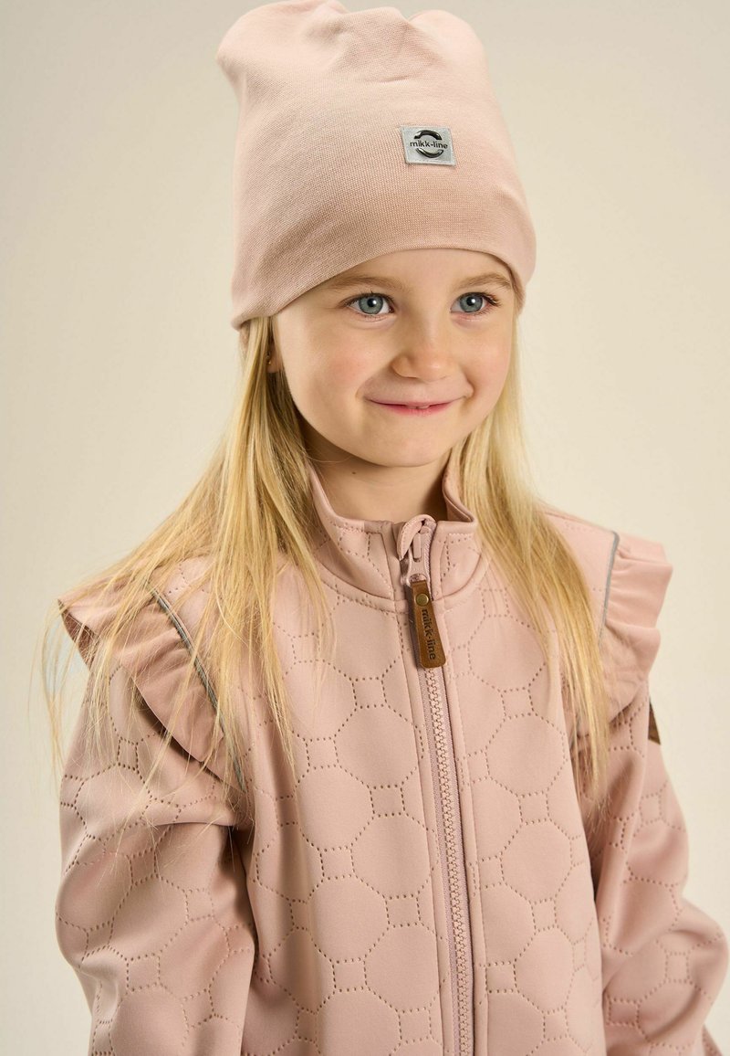 Jeune fille aux longs cheveux blonds portant un bonnet rose pâle et une veste matelassée assortie avec des épaules volantées, souriant légèrement.