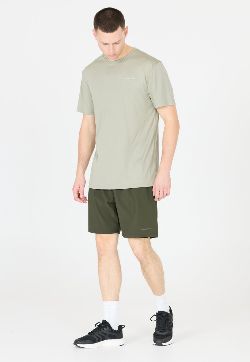 Licht olijfgroen t-shirt met korte mouwen, gecombineerd met donkergroene shorts. Zwarte sportschoenen met witte sokken. Eenvoudig, functioneel ontwerp.