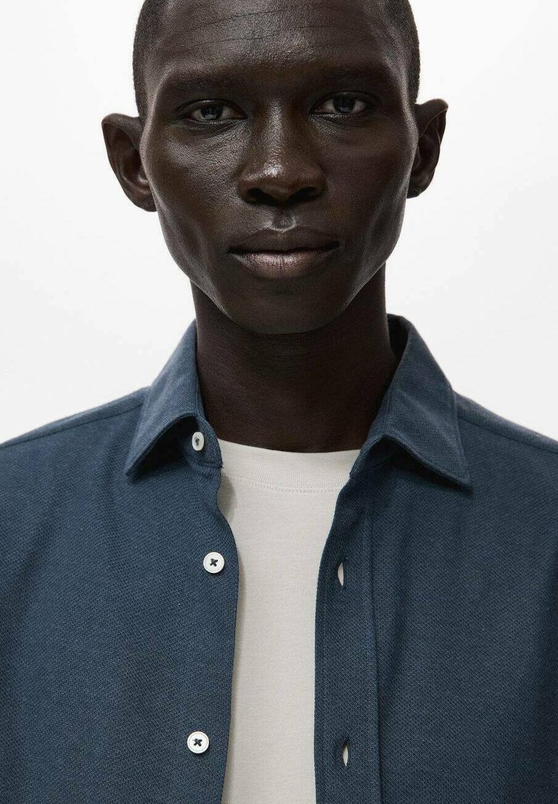 Mørkeblå tekstureret skjorte med knap-op design, der har hvide knapper og en lys beige skjorte under. Enkel krave detaljer.