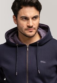 Granatowa bluza z kapturem na zamek, wykonana z mieszanki bawełny, z kontrastującą fioletową podszewką, ozdobiona naszywką z logo i akcentami ze sznurka na kapturze.