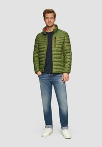 Grüne Daunenjacke mit gestepptem Design, hohem Kragen und einer Reißverschlusstasche. Trägt man über einem marineblauen Hemd und kombiniert mit hellblauen Jeans und weißen Sneakern.