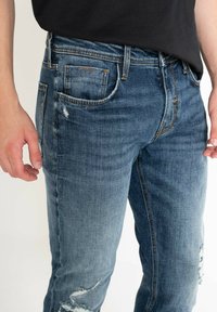 Jeans de mezclilla azul desgastados con un corte ajustado, que presentan parches desgastados, bolsillos estándar y detalles de costura en naranja contrastante.