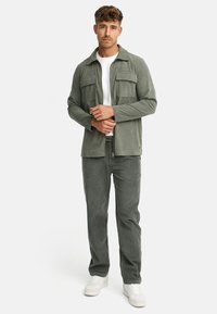 Green corduroy jacket with two chest pockets, paired with matching trousers and white sneakers. Smooth texture and relaxed fit.