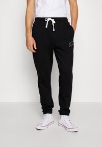 Pantalones deportivos negros con cintura elástica, cordón blanco, bolsillos laterales y detalle de logo bordado en el muslo izquierdo.