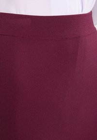 Detail osoby v bílém topu zastrčeném do vysokého pasu žebrované tmavě fialové sukně nebo kalhot.