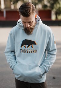 Felpa leggera di colore blu chiaro, realizzata in tessuto morbido, con una grafica di un orso nero e il testo "FORSBERG" in grassetto. Include una tasca a marsupio frontale.