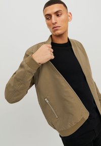 Khaki bomber jacket with ribbed cuffs and waist, featuring a zippered side pocket and a smooth, matte texture. Worn over a black sweater.