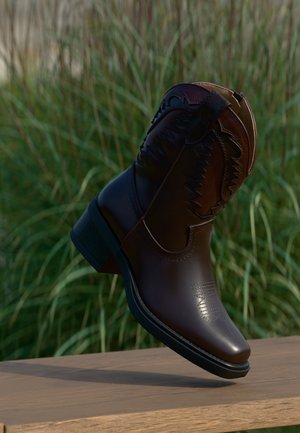 Cizmă de cowboy din piele maro închis cu broderie decorativă, poziționată înclinat pe o suprafață de lemn, cu fundal de iarbă înaltă.