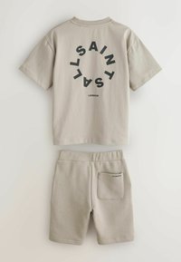 Bež bombažni komplet majice in kratkih hlač. Majica ima črni okrogli logotip z napisom "SAINTS" in "LONDON." Kratke hlače imajo zadnji žep z oznako blagovne znamke.