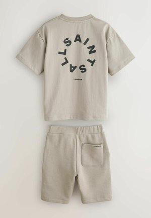 Beżowy zestaw z bawełnianą koszulką i szortami. Koszulka ma czarne okrągłe logo z napisem „SAINTS” i „LONDON”. Szorty posiadają tylną kieszeń z brandingiem.