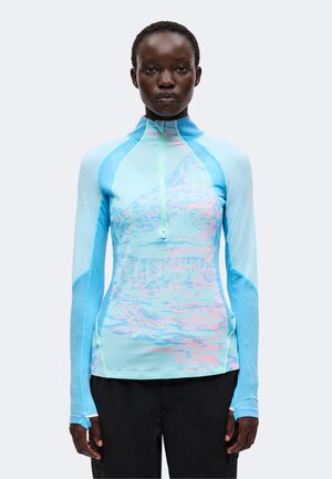Femme portant un haut à manches longues bleu clair avec un motif montagne et une fermeture éclair à l'avant, debout devant un fond uni.