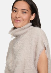 Junge Frau mit dunklem Haar trägt einen hellbeigen, flauschigen ärmellosen Pullover mit hohem Kragen und Reißverschluss und lächelt leicht vor einem weißen Hintergrund.