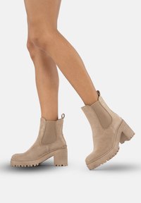 Stivaletti alla caviglia in suede beige con tacco spesso, pannelli laterali elastici e un tab di loop sul retro, dotati di una suola in gomma testurizzata.