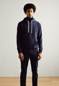 Sudadera con capucha de color azul marino con cordones blancos, bolsillo frontal y bajo de canalé, combinada con pantalones ajustados a juego. Textura suave.