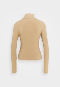 Top beige à manches longues avec un col haut, texture côtelée, coupe slim, et une petite étiquette de marque au dos, conçu pour une silhouette ajustée.