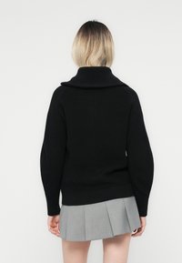 Zwart gebreide trui met een hoge kraag, voorzien van brede mouwen en ribbeltextuur, gecombineerd met een grijze geplooide rok.