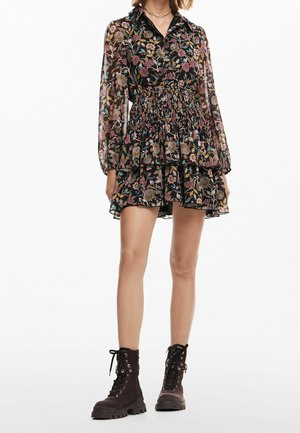 Femme portant une robe noire à fleurs à manches longues avec une taille smockée et un ourlet volanté, associée à des bottes de combat noires à lacets.