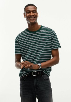 Hombre sonriente que lleva una camiseta de rayas verdes y blancas, vaqueros negros, cinturón negro y reloj de pulsera plateado, de pie con las manos juntas.