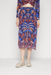 Jupe crayon tricotée en bleu avec des motifs de palmiers et de fleurs vibrants, présentant des franges à l'ourlet. Associée à des loafers plateforme beiges.