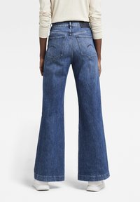 Breda ben blå denimjeans med medelhögt midjeområde, utrustad med två bakfickor och en ren fåll. Tyget har en lätt textur och vintageutseende.